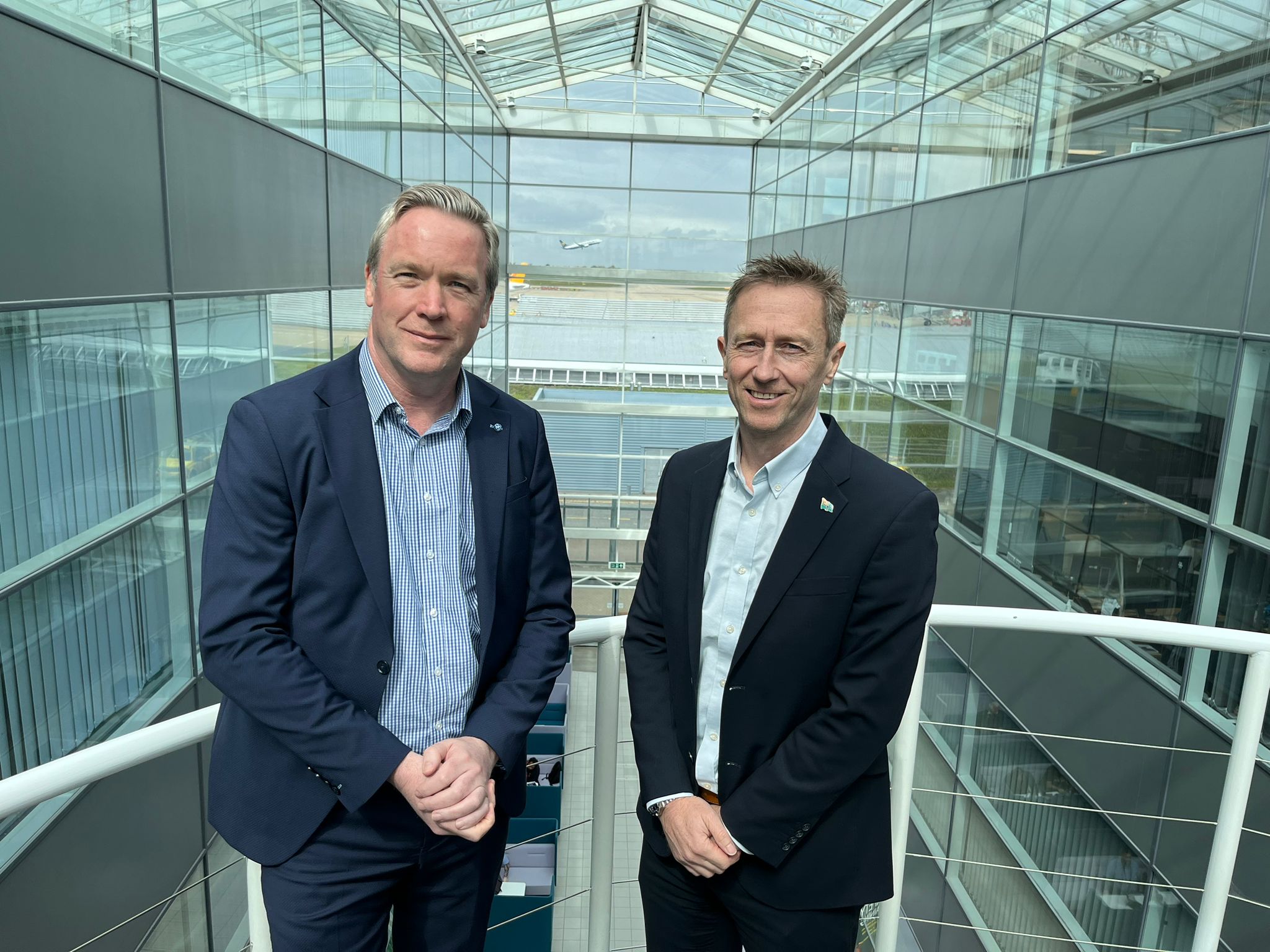 Ken O'Toole and Rob Bishton standing together in a glass entryway overlooking a plane taking off in the distance at Stansted airport.