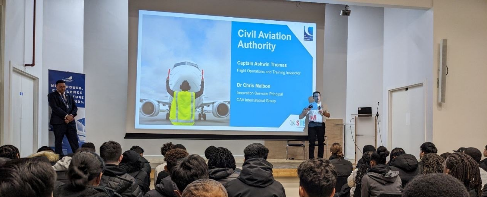 Image of students listening to presentation by two speakers with projector board