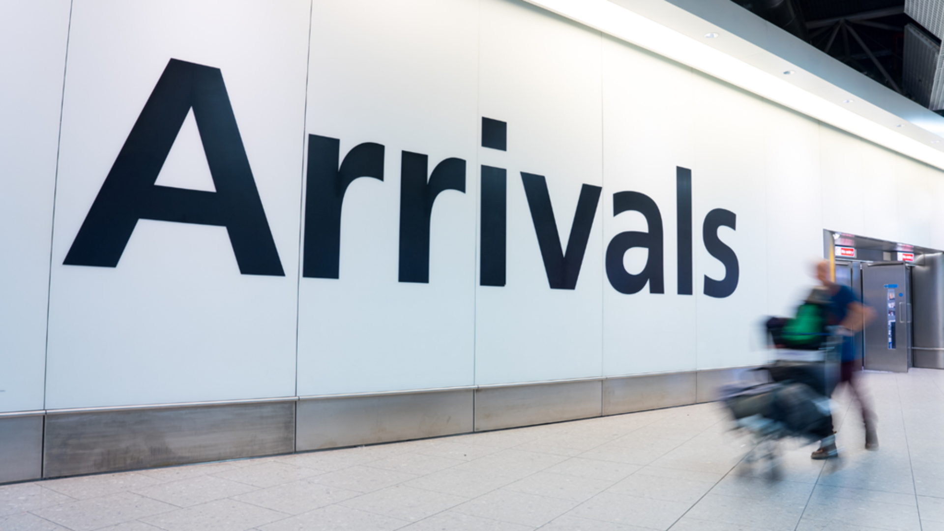 An image of an arrivals sign at an airport with a person blured with luggage walking past the sign. 