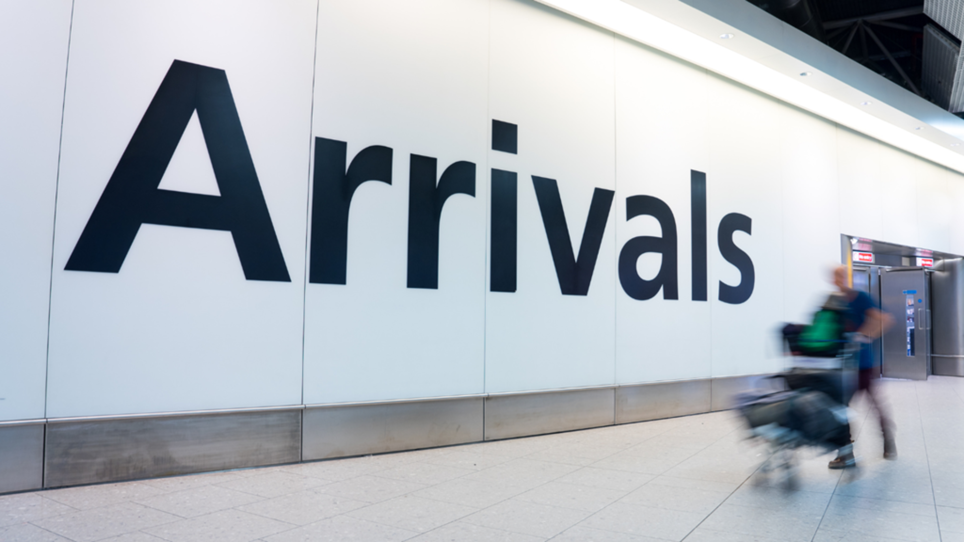 An image of an arrivals sign at an airport with a person blured with luggage walking past the sign.