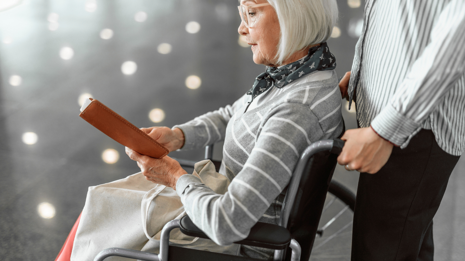 An image of an elderly lady in a wheelchair and another person pushing the wheelchair